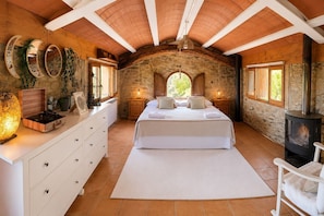 A bedroom with a stone wall, wooden ceiling beams, a bed, a dresser, a record player, and a wood-burning stove.