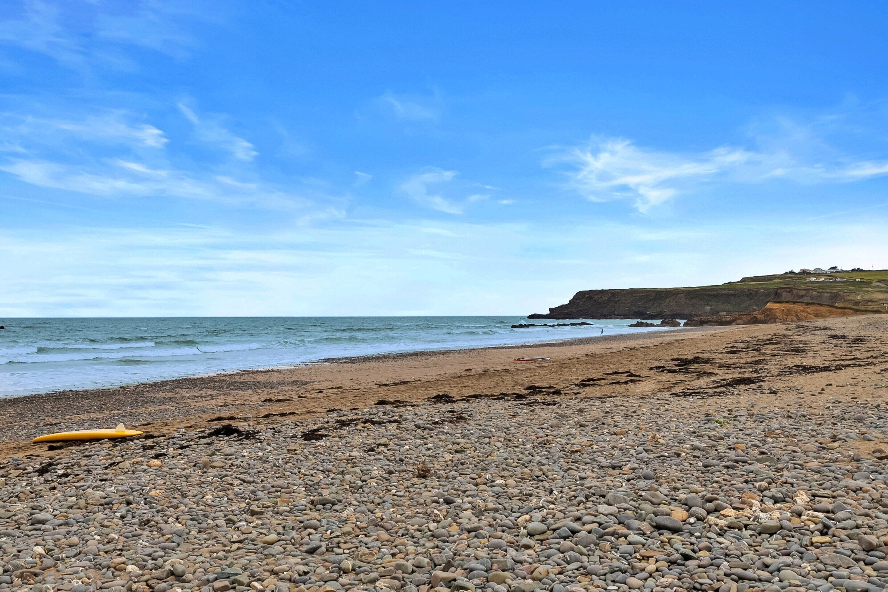 A pebble beach with a surfboard, calm sea, and a cliff in the background.