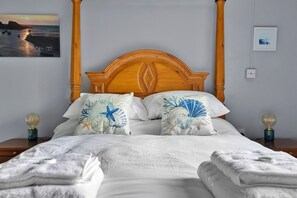 Una cama con cabecera de madera, dos almohadas decorativas con motivos marinos, una colcha blanca y dos lámparas de noche con pantallas de cristal esférico.