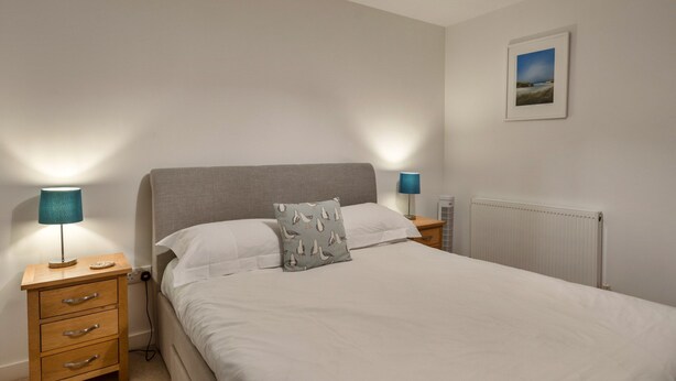 A neatly made bed with a pillow, two bedside tables with lamps, a radiator, and a framed picture on the wall. - Stunning Broad Haven Beach House (Newquay)