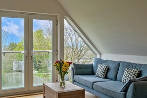A room with a blue sofa, a wooden coffee table, and a vase of flowers. There are large glass doors leading to an outdoor area with trees. - Stunning Broad Haven Beach House (Newquay)