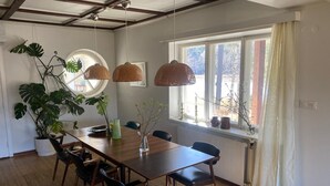 A dining area with a wooden table, chairs, hanging rattan lamps, potted plants, and a window with curtains.