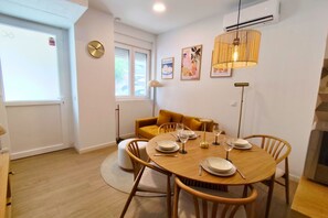 En matplats med ett runt träbord för fyra personer, omgiven av trästolar. I bakgrunden finns en soffa med gula kuddar, en golvlampa och en väggmonterad luftkonditioneringsanläggning.