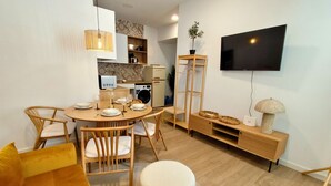 A modern living room with a round dining table, wooden chairs, a sofa, a television mounted on the wall, and a built-in washing machine. - Travel Habitat Marchalenes Apartment - 3 BedRooms (Valencia)
