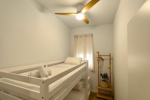 Ein Zimmer mit einem Einzelbett, einem Deckenventilator, einem Fenster mit Vorhängen und einem Holz-Kleiderständer mit einer Topfpflanze.