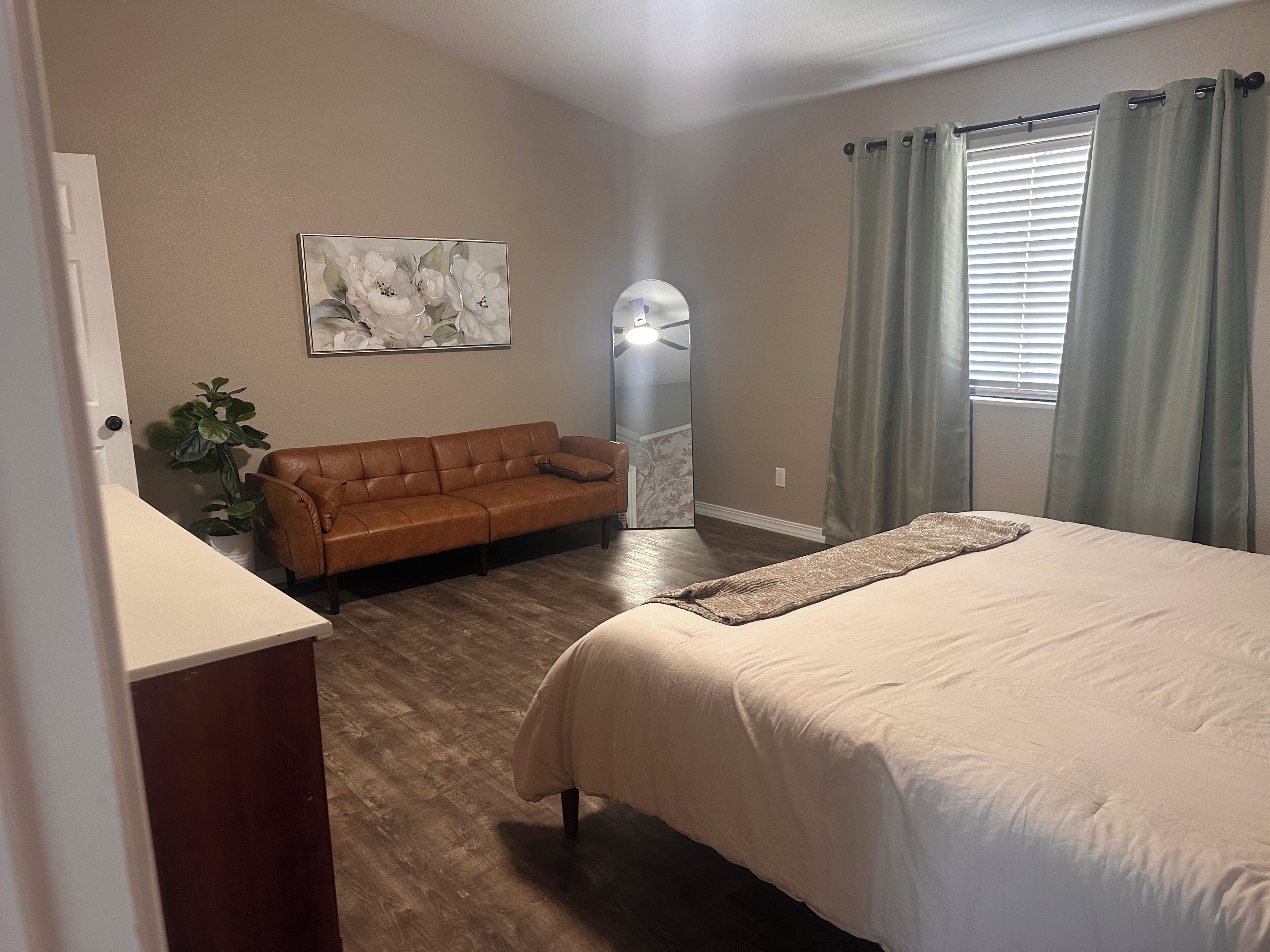 A bedroom with a bed, a brown leather sofa, a potted plant, and a wall-mounted artwork.