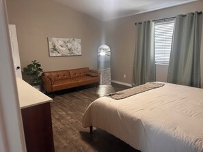 A bedroom with a bed, a brown leather sofa, a potted plant, and a wall-mounted artwork.