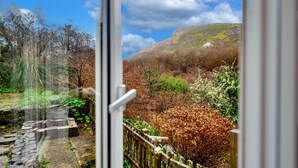 A view through a window of a stone pathway leading to a wooden fence, with a hill and trees in the background. - Historic Cornish Valley Cottage for 5 (Bude)