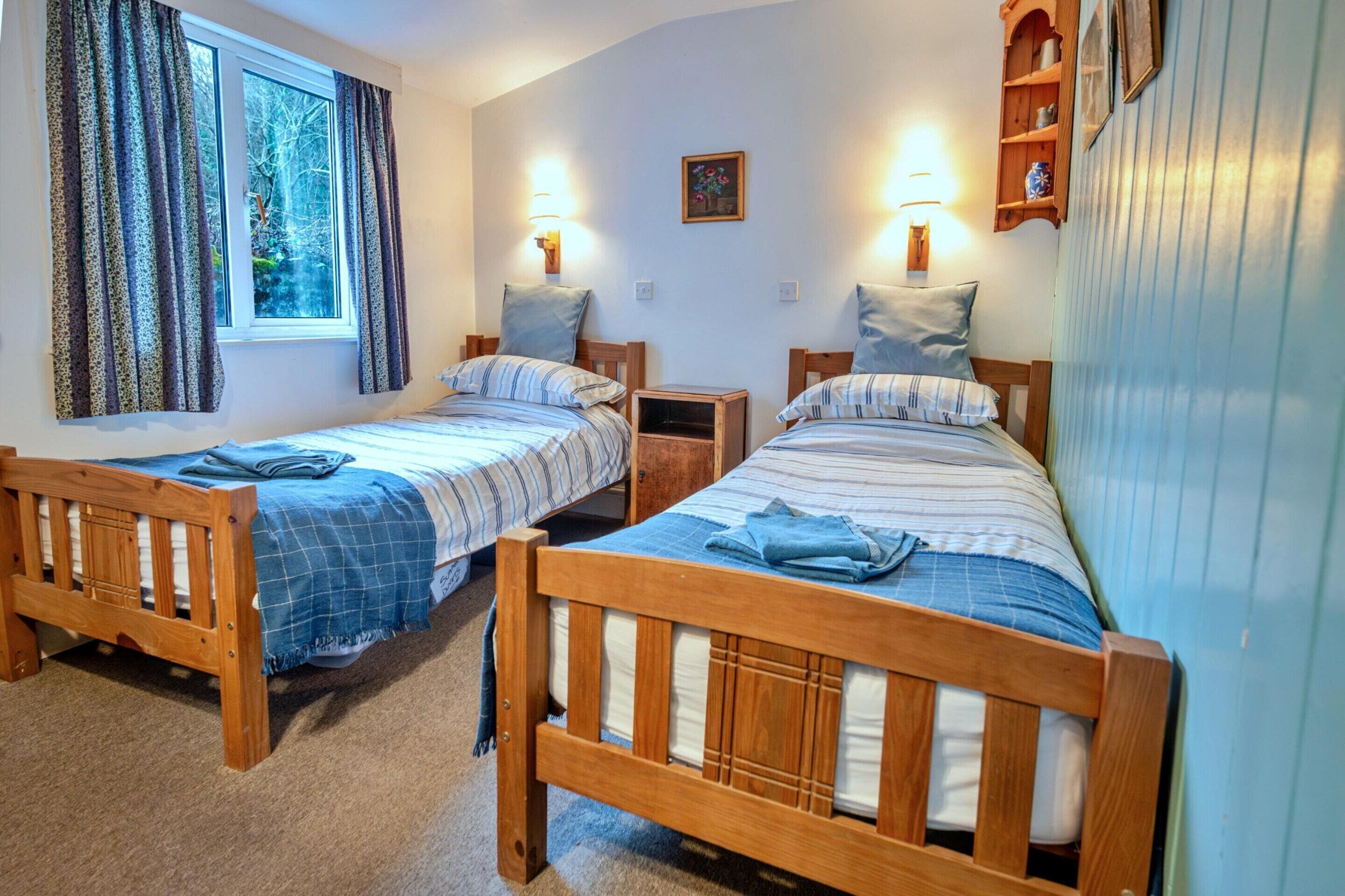 A room with two wooden beds, each with a blue blanket and pillow, a small bedside table, and a window with curtains.