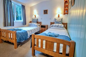 A room with two wooden beds, each with a blue blanket and pillow, a small bedside table, and a window with curtains. - Historic Cornish Valley Cottage for 5 (Bude)