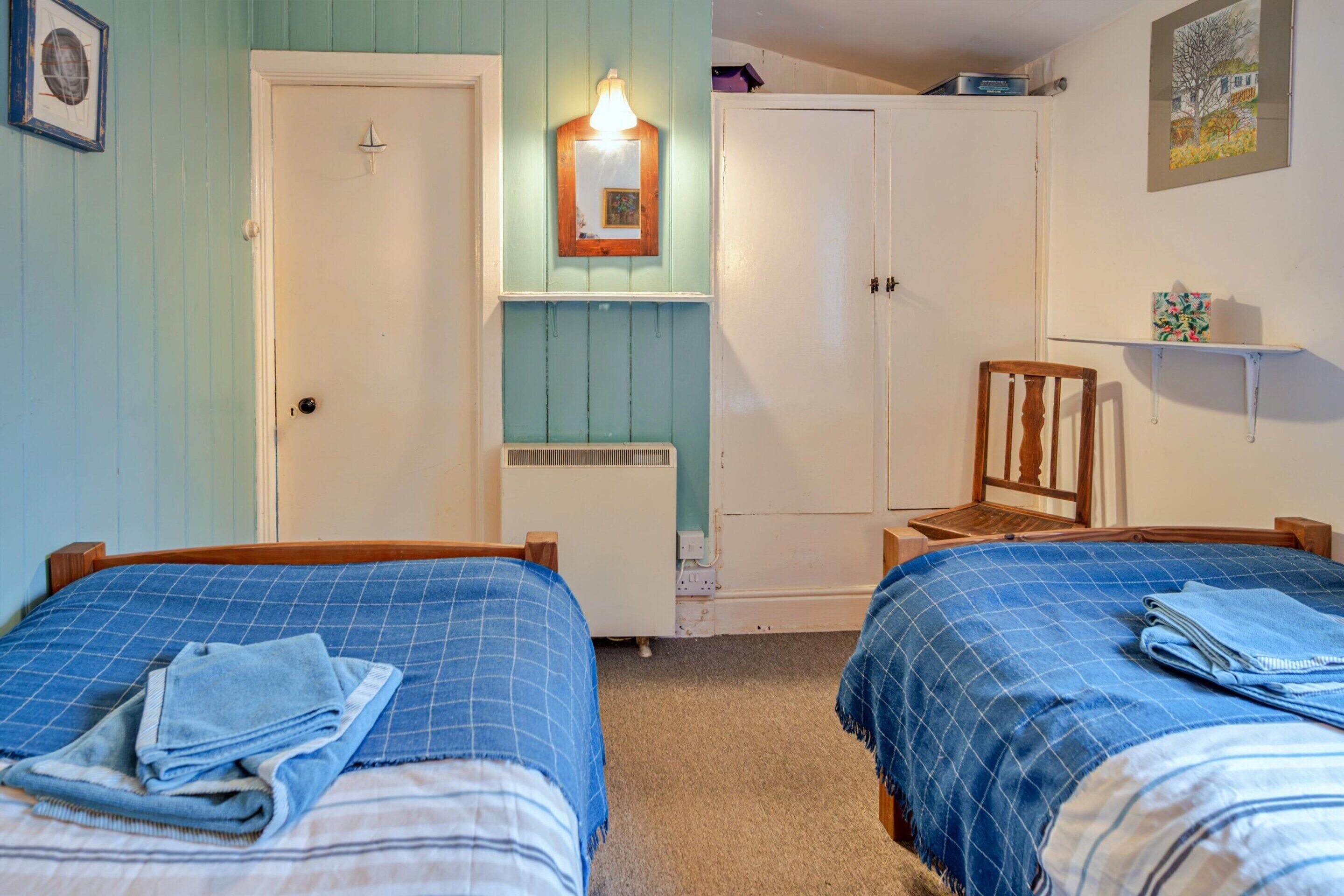 A bedroom with two beds, a chair, a wardrobe, and a shelf with a painting.