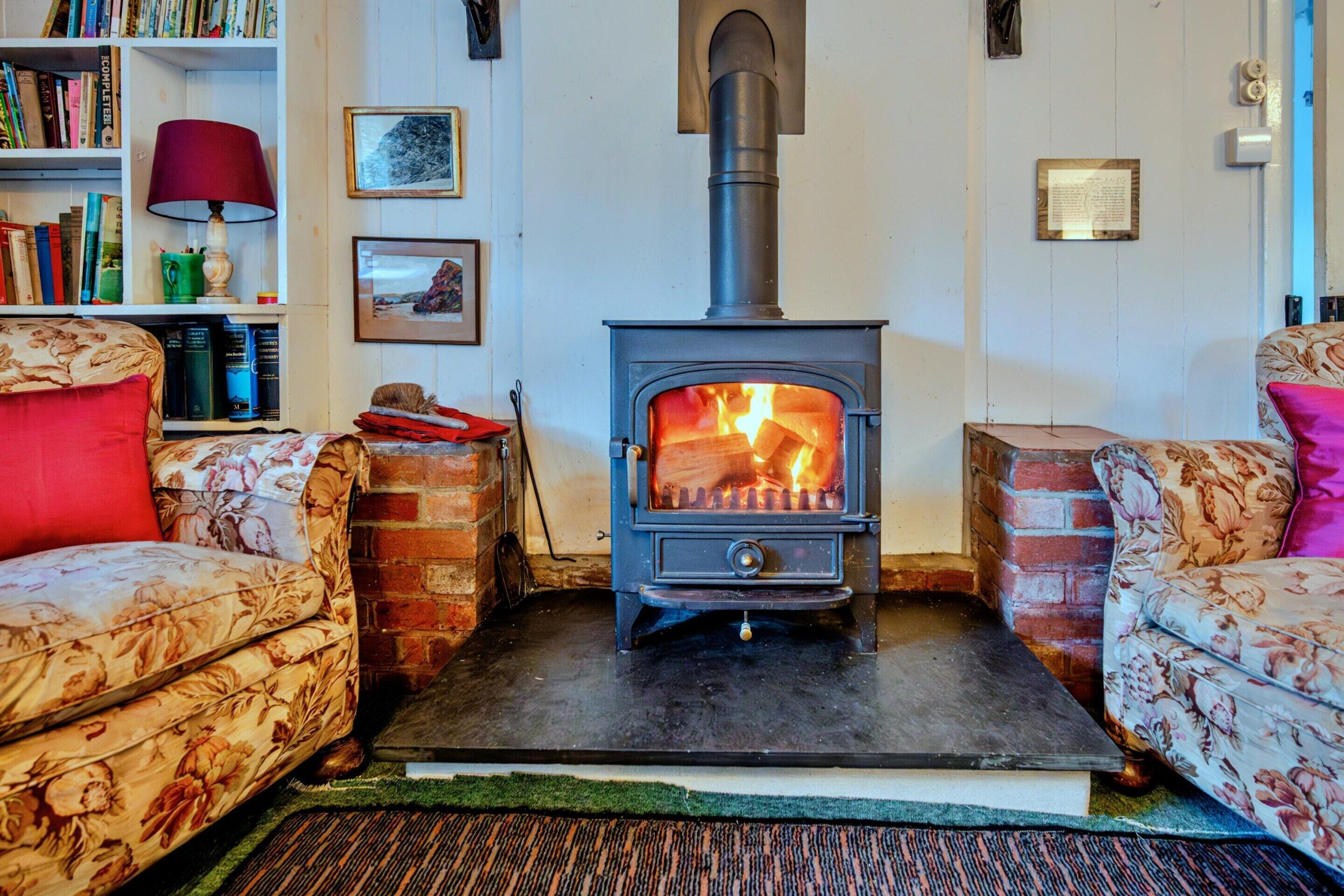 A cozy living room with a wood-burning stove, floral-patterned sofas, a bookshelf, and a red lamp.