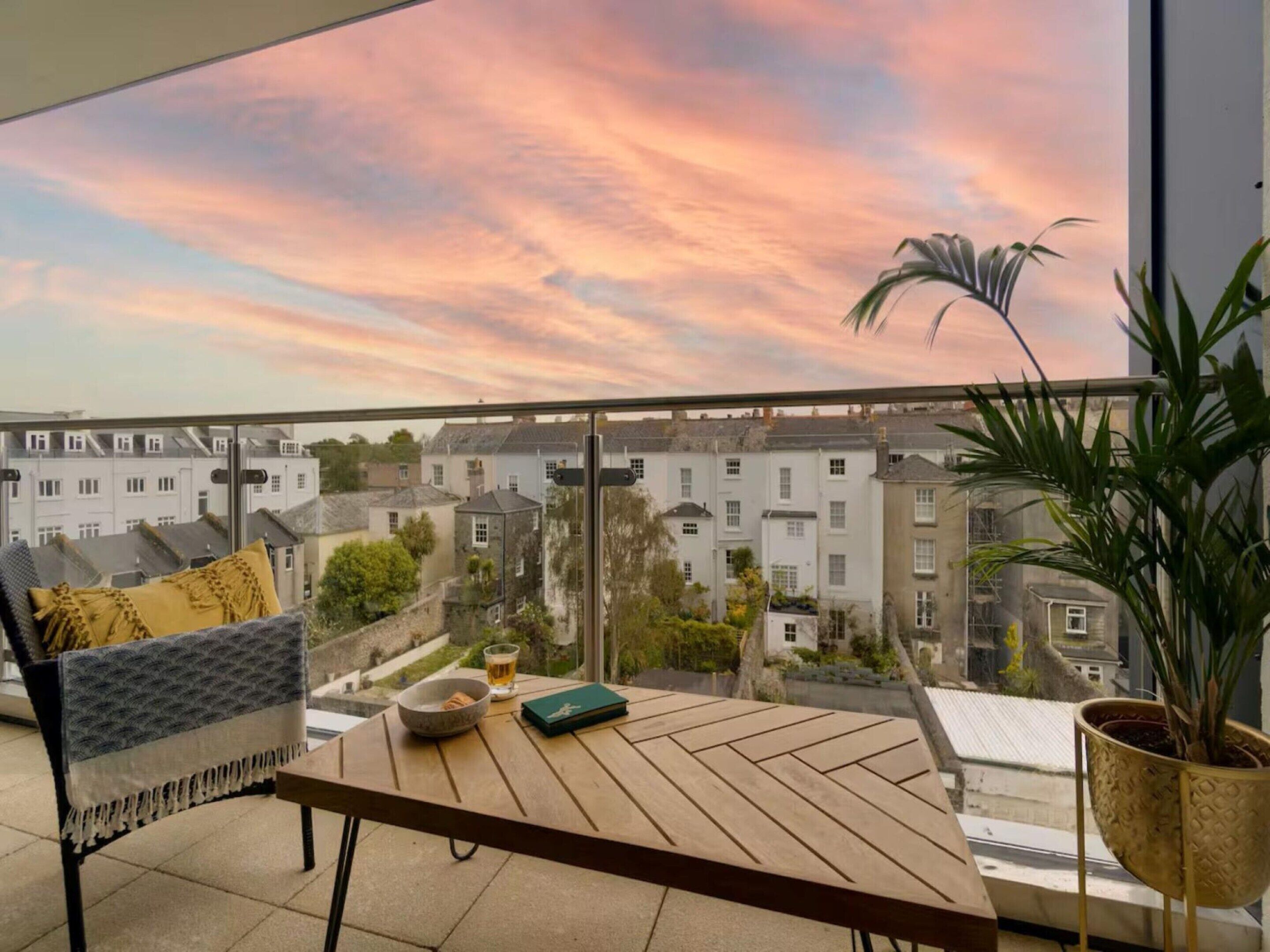 Un balcon donnant sur des immeubles d’habitation, une table avec un bol et un livre, et une plante en pot.