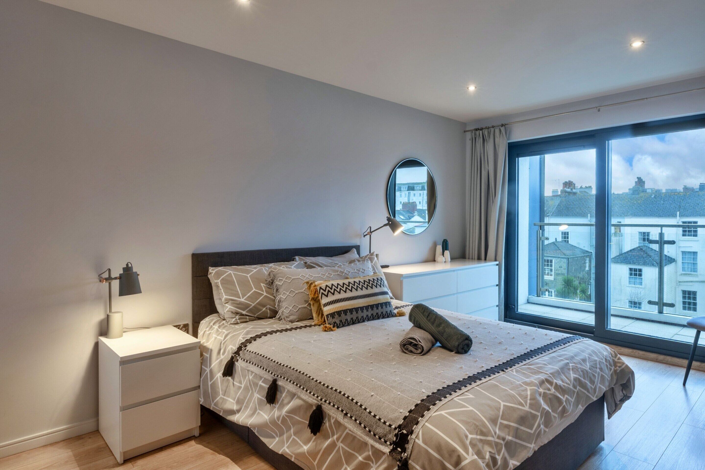 A bedroom with a large bed, a nightstand, a bedside lamp, a mirror, and a view of buildings through a large window.