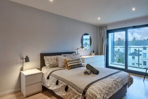 A bedroom with a large bed, a nightstand, a bedside lamp, a mirror, and a view of buildings through a large window. - Modern Plymouth Apartment Near The Hoe (Plymouth)