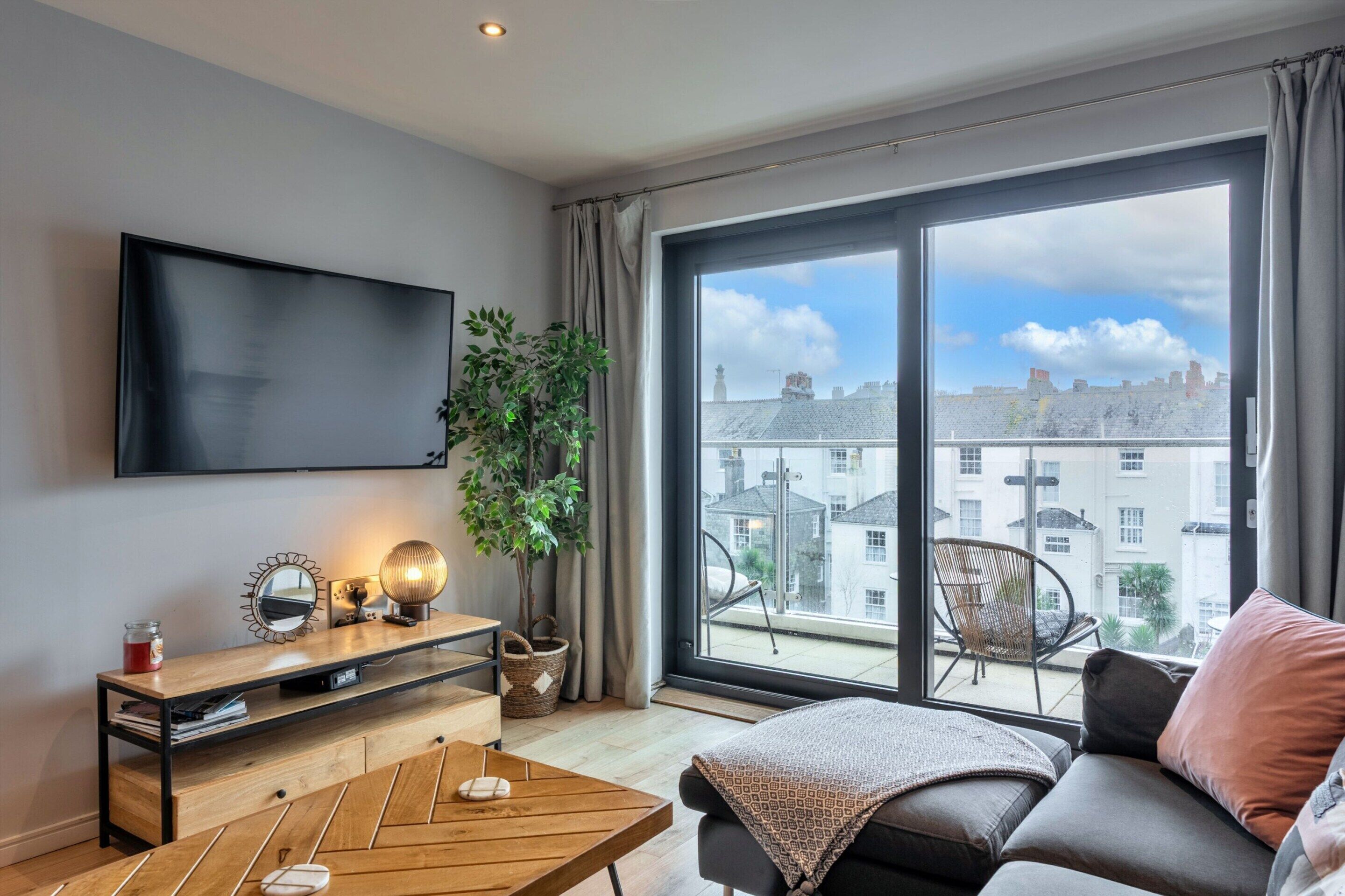 Un salon moderne avec un téléviseur à écran plat, une table basse en bois, un canapé agrémenté d’un plaid, une plante en pot et une vue sur le paysage urbain à travers de grandes fenêtres.