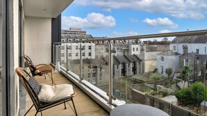 A balcony with a view of residential buildings, a chair, a cushion, and a glass railing. - Modern Plymouth Apartment Near The Hoe (Plymouth)