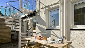 Una mesa de madera al aire libre con alimentos para el desayuno, una silla de metal y una escalera que lleva a un balcón.