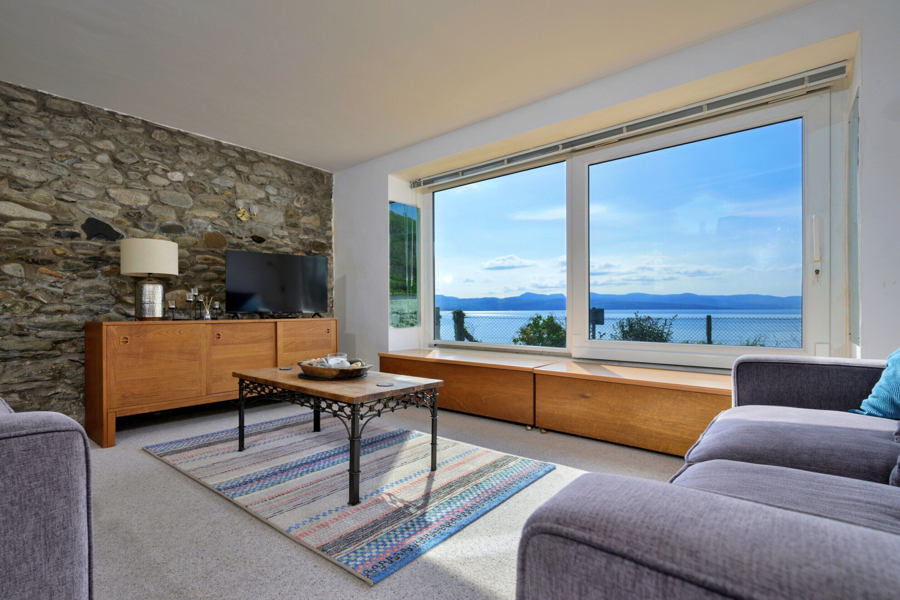 A modern living room with a stone wall, a wooden cabinet, a television, and a large window with a view of the sea.