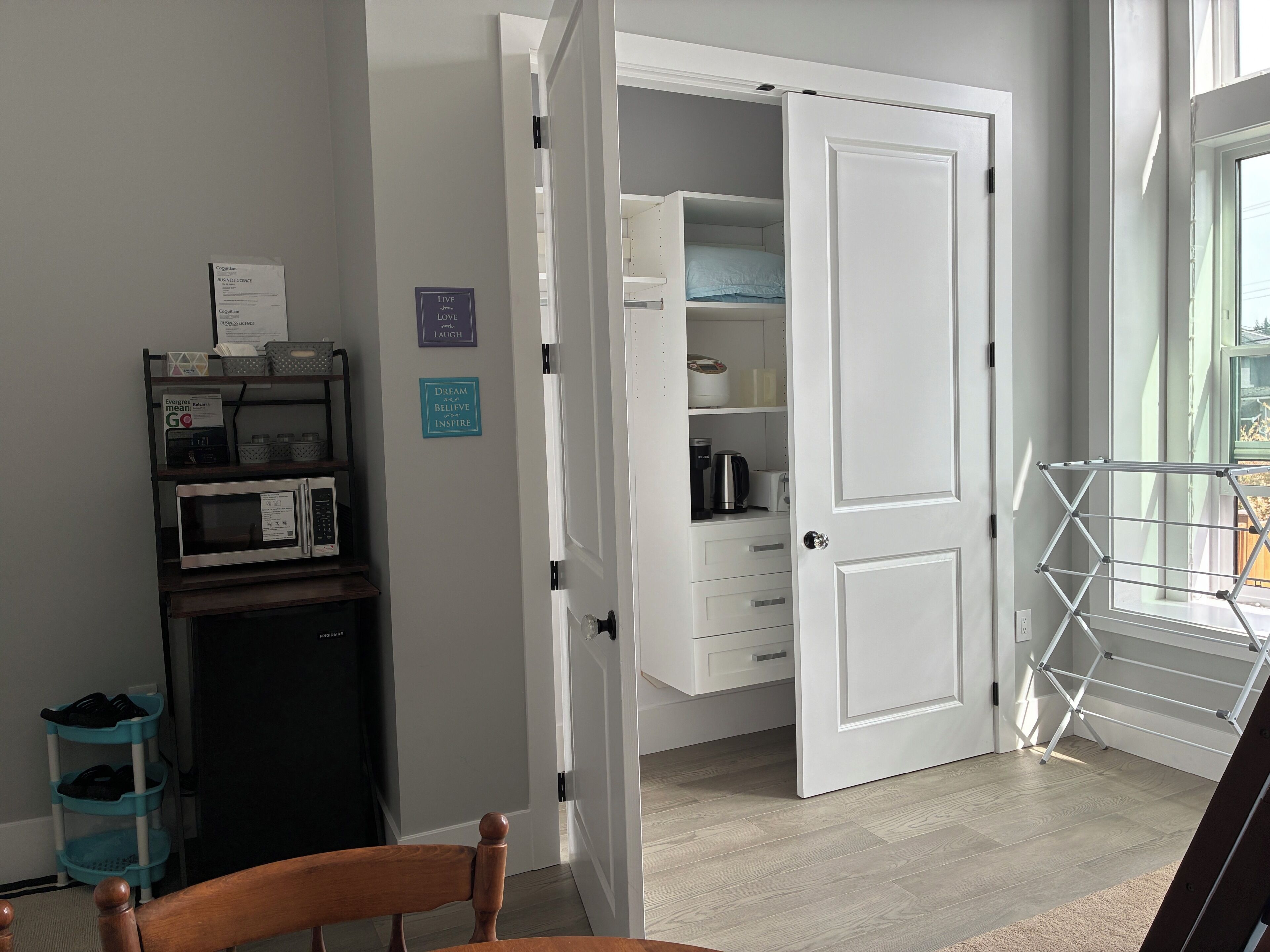 A room with a white door, a microwave, a shelf with storage bins, a chair, and a window with a view.