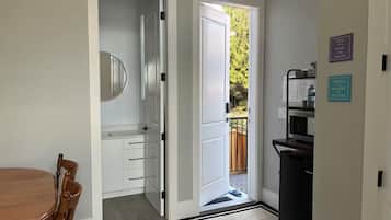 Ein Zimmer mit einem Esstisch, Stühlen, einem weißen Schrank, einer schwarzen Mülltonne und einer Tür, die zu einem Außenbereich mit einer Holzterrasse führt.