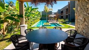 Zona junto a la piscina con una mesa redonda y sillas, rodeada de exuberante vegetación y un muro de piedra.