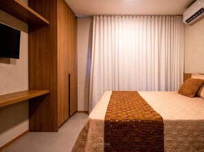 A hotel room with a bed, a TV mounted on the wall, a wardrobe, and an air conditioning unit.