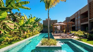 Una piscina con sombrilla y sillas de descanso, rodeada de exuberante vegetación tropical y un edificio moderno.