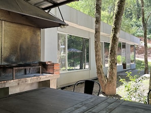 A modern outdoor patio with a brick fireplace, large windows, and a view of the surrounding forest.