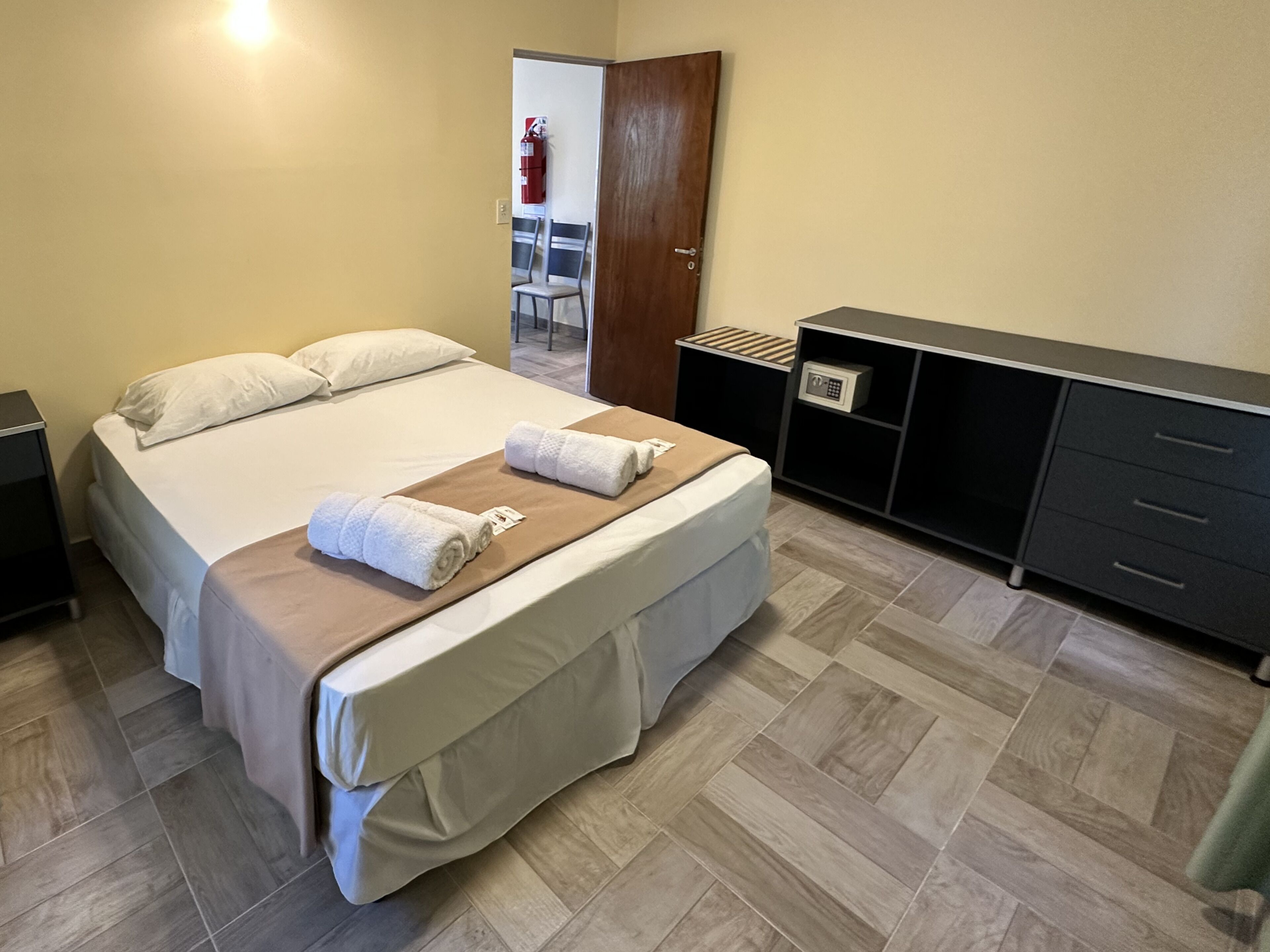 Een hotelkamer met een bed, nachtkastje, dressoir en een brandblusser aan de muur.