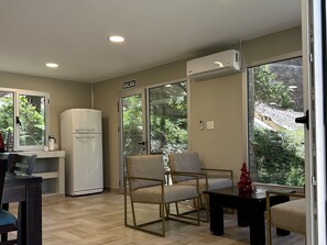 A room with a kitchenette, a sofa, a chair, and a table. There is a window with a view of greenery outside.