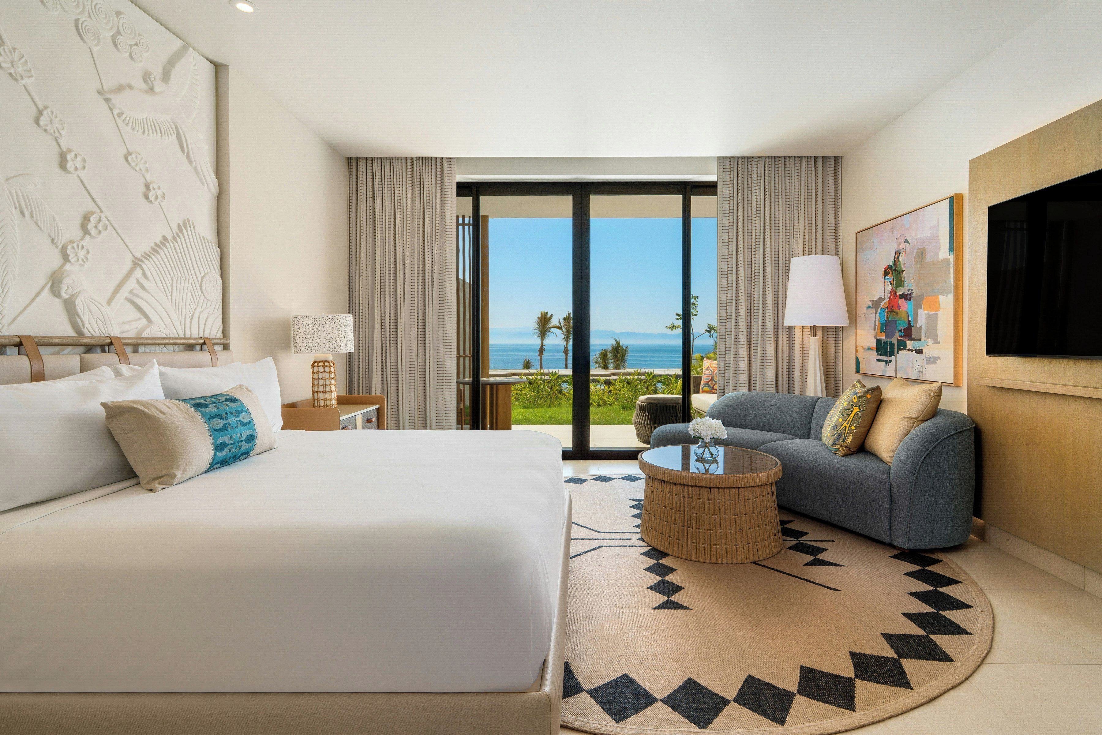 A hotel room with a bed, a sofa, a coffee table, a TV, and a view of the ocean.