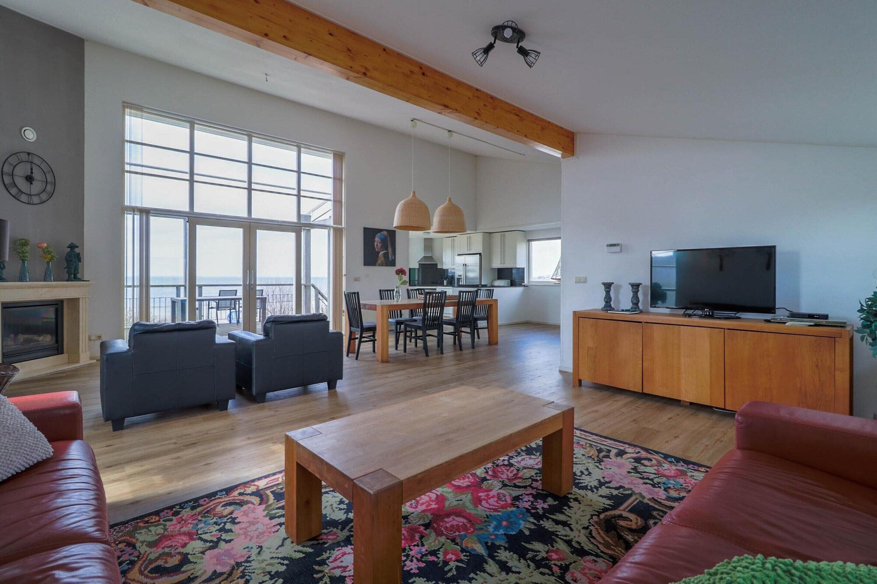 A spacious living area with a wooden ceiling, a dining table with chairs, a television on a wooden cabinet, and a large window providing natural light.