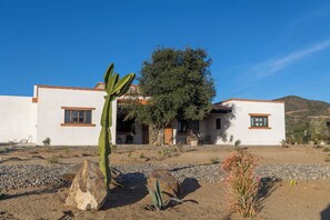 Un bâtiment à deux étages, doté d’une façade blanche et de bordures marron, entouré de végétation désertique et surplombé d’un ciel bleu clair.