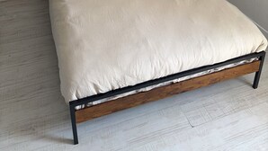 A single bed with a wooden frame and a headboard, a beige bedspread, and two dark pillows.