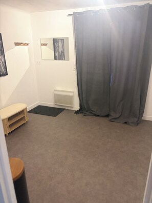 Une pièce dont le sol est recouvert d’une moquette, qui contient un banc, un petit radiateur, un miroir et un rideau.