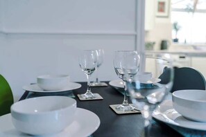 Taula de menjador preparada amb plats blancs, bols i copes de vi, amb vista a una cuina al fons.