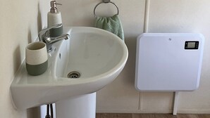 Un baño con un lavamanos blanco, un dispensador de jabón, un perchero con toallas verdes y un calentador eléctrico blanco.