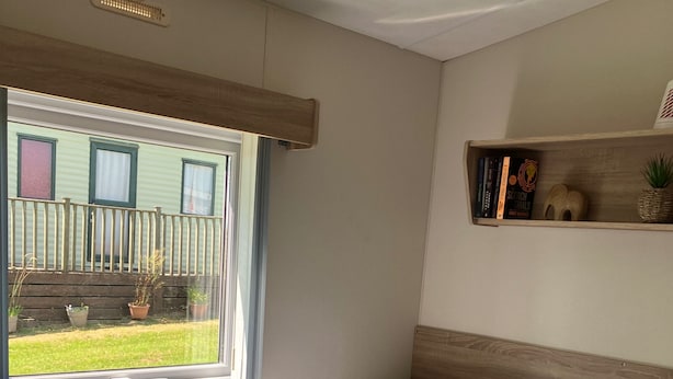 Una habitación con dos camas, una ventana con cortinas, una mesita de noche con lámpara, y un estante con libros y una planta.