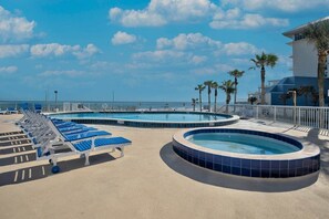 Een zwembadgedeelte met ligbedden en een jacuzzi, omgeven door een hek en palmbomen, met een helder uitzicht op de oceaan.