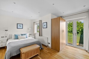 Ein Schlafzimmer mit Bett, Nachttischen, einer Bank, einem Heizkörper, einem Fenster mit Vorhängen und einer Tür, die auf einen Balkon führt.
