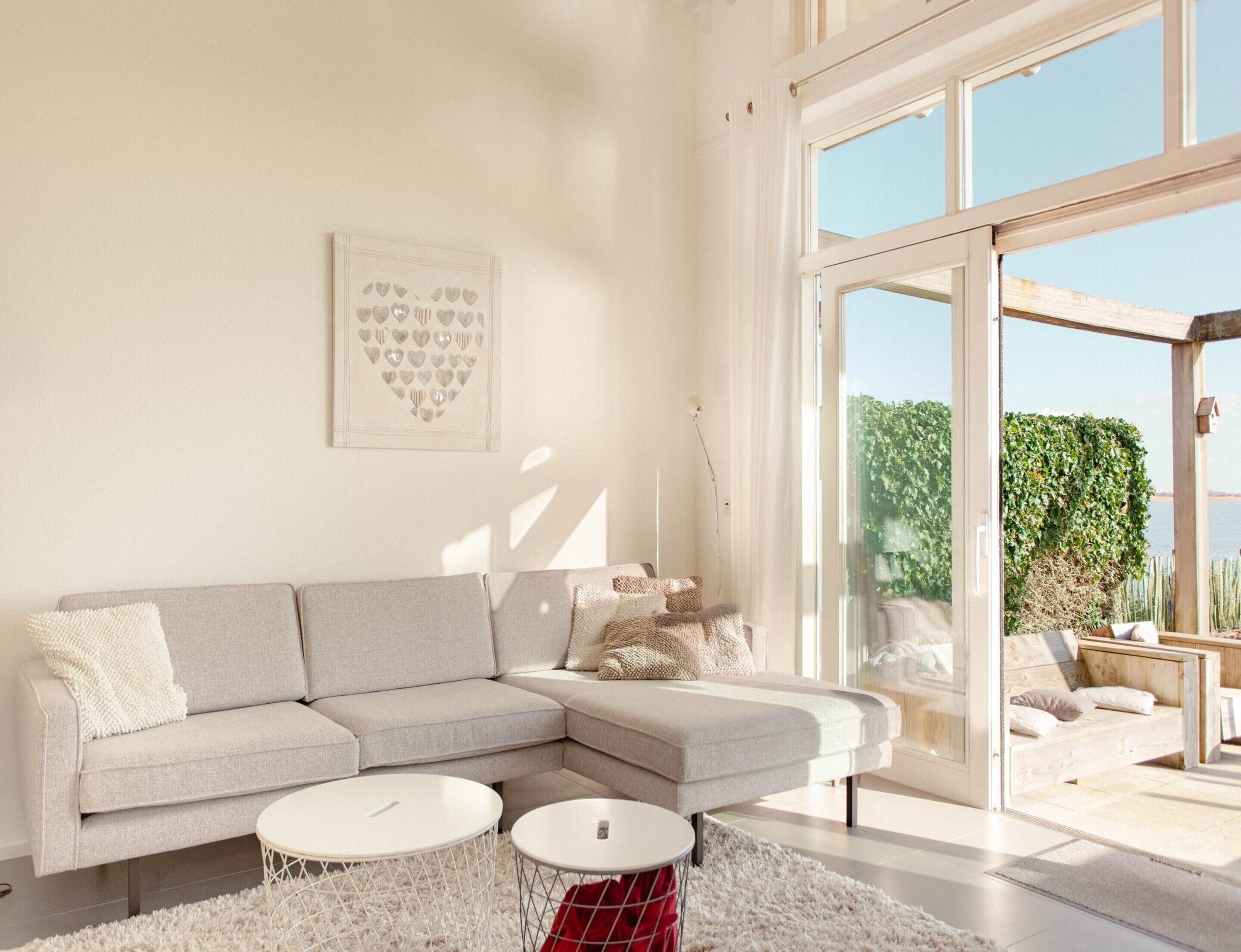 Un salon moderne avec un canapé d’angle, deux petites tables basses rondes blanches et une grande porte vitrée donnant sur un espace extérieur.