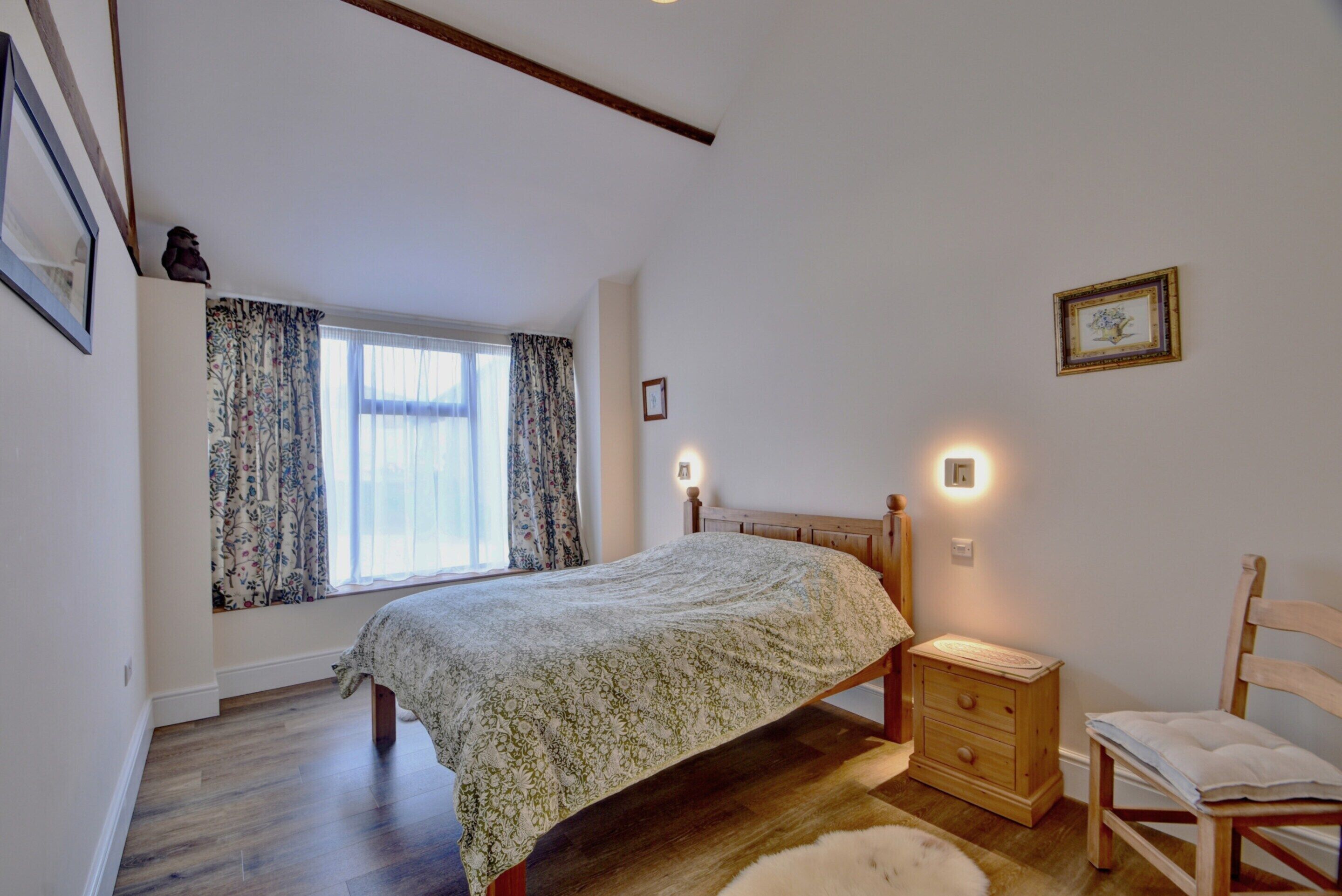 A bedroom with a bed, a nightstand, a chair, a window with curtains, and a wall-mounted light.