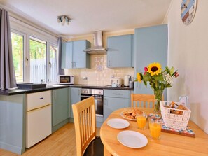 A compact kitchen with a microwave, oven, and sink. There is a dining table with plates, glasses, and a basket of bread. - Cosy Cornwall Bungalow for 4 (St Ives)