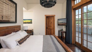 Ein Hotelzimmer mit einem großen Bett, einem Holzkopfende, einem Bild an der Wand, einer Tür, einem kleinen Tisch mit Lampe und Blick durch große Fenster.