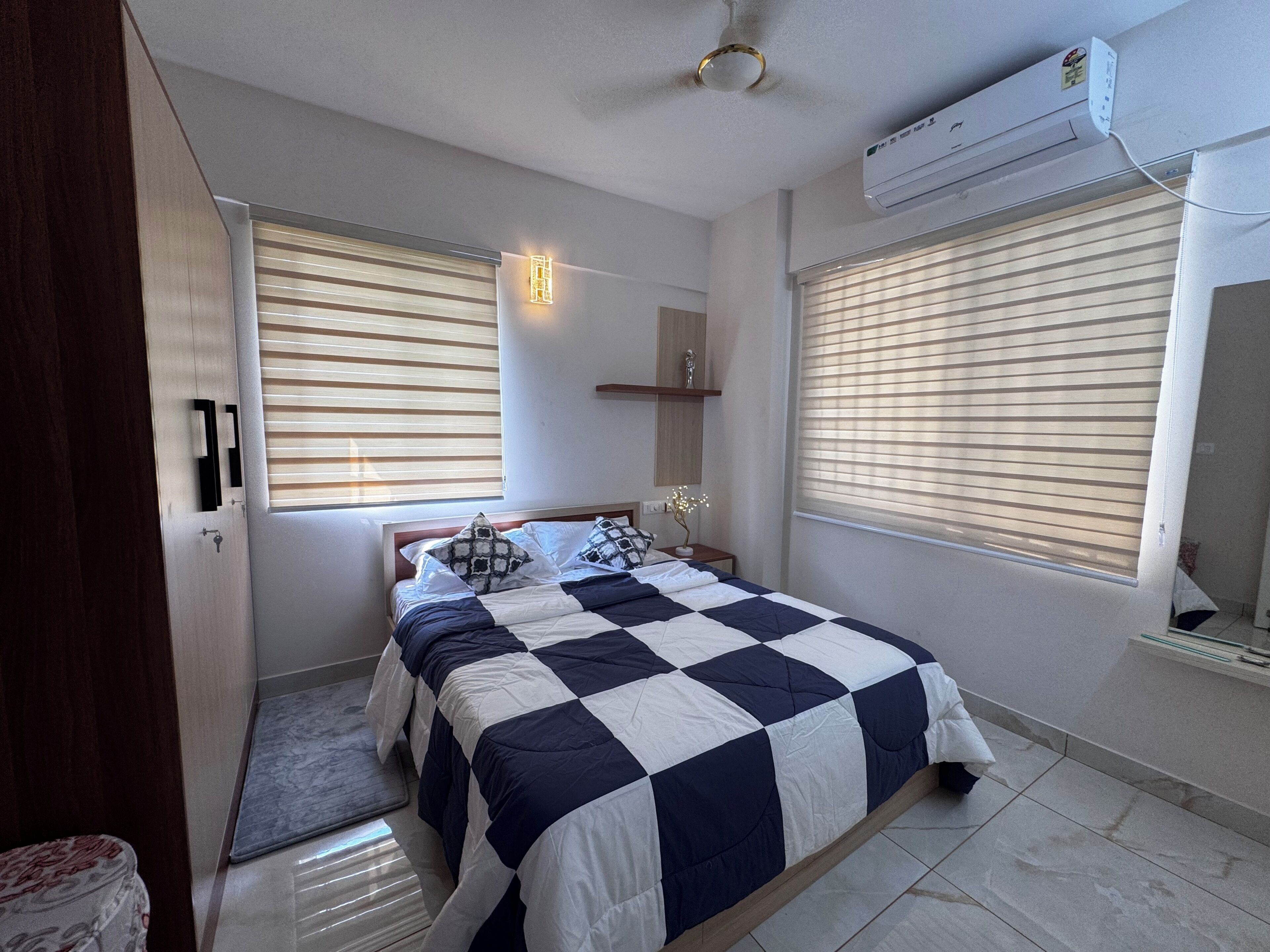 A bedroom with a bed, a bedside table, a nightstand, a wall-mounted shelf, and a window with blinds.