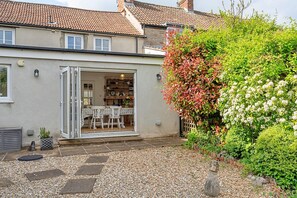 Ein Haus mit Terrasse, ein Garten mit verschiedenen Pflanzen und ein sichtbarer Innenraum mit Esstisch und Stühlen.