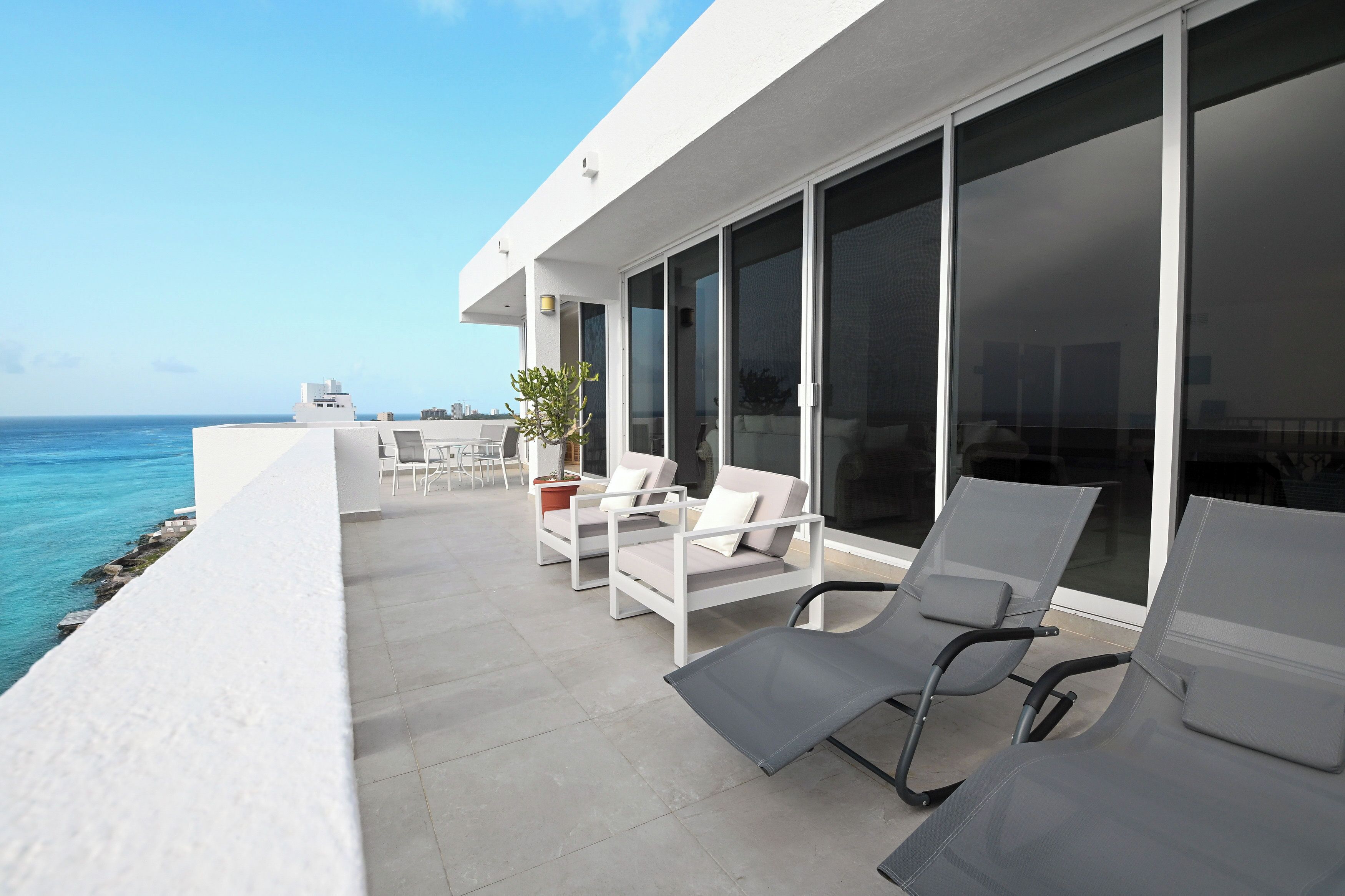 Un balcon extérieur moderne offrant une vue sur l’océan, aménagé avec des chaises et une table blanches.
