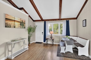 A room with a wooden floor, a white bench, a table with a lamp, a door, and a window with curtains.