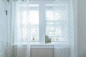 Ein Zimmer mit Fenster, weißen Jalousien und einem transparenten Vorhang.